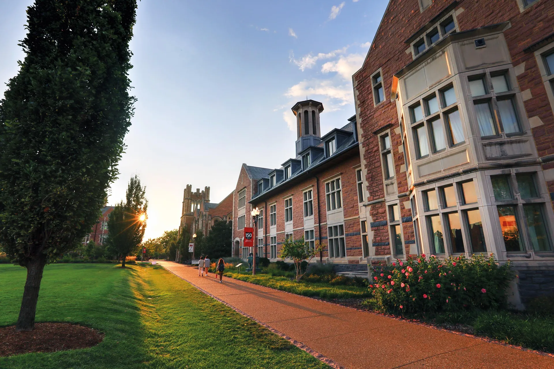 Knight Center building with large windows and red stone facade at sunset, with a few people walking along a path and sunlight filtering through trees.